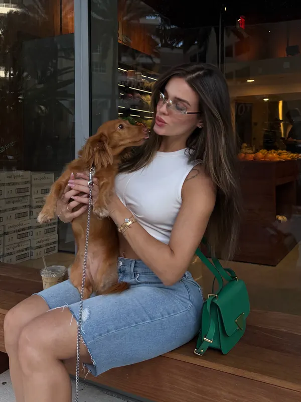 Gabryelle Pedroso seated at a cafe holding a small dog while wearing a white top and denim skirt